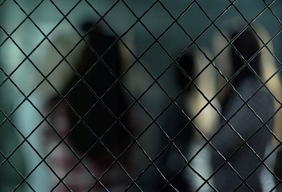 A wire mesh obscures the view of figures standing in what could be a prison. The image is associated with the topic of sexual abuse in prisons.